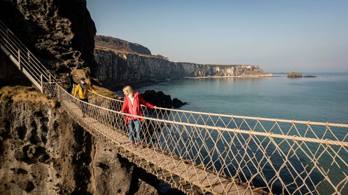 Two visitors walking along Carrick-a-Rede Rope Bridge. Visitor at the front looking down to the Atlantic Ocean. Visitor at the back looking ahead to the island.